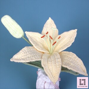 May include: A crocheted cream-colored lily flower with a matching closed bud, green stems, and orange-tipped stamens. The flower is set in a white crocheted base. A small square logo in the corner displays the text Liz Crochet Gifts.