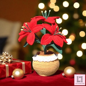 May include: A handmade crochet poinsettia plant with vibrant red petals and green leaves, displayed in a beige pot with a white lace trim. The poinsettia is set on a red surface with a wrapped gift and gold ornaments.