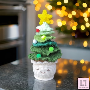 May include: A crocheted Christmas tree with a yellow star on top, green and white layers, and a white pot with a smiling face. The tree is sitting on a table with a blurred background of a Christmas tree.