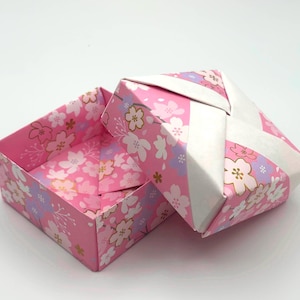 May include: A pink origami box with a lid, decorated with a floral pattern of white, pink, and lavender blossoms. The box is open, revealing the interior with the same floral design.