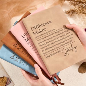 May include: A stack of notebooks in various colors, including tan, pink, brown, and blue. The top notebook is tan and features the text "Difference Maker" with a definition and the name "Stanley".