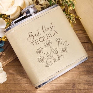Könnte beinhalten: Eine beigefarbene Flachmann aus Kunstleder mit silbernem Verschluss. Der Flachmann ist mit dem Schriftzug "But first TEQUILA" und einem Blumendesign verziert. Der Flachmann ist rechteckig und hat einen genähten Rand.