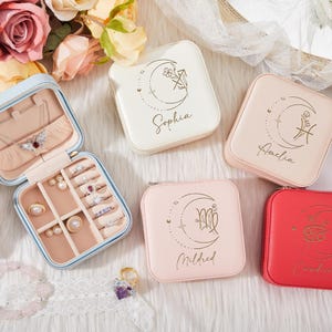 Könnte beinhalten: Eine offene hellblaue Schmuckschatulle mit Ringen, Halsketten und Ohrringen. Vier geschlossene quadratische Schmuckkästchen in Creme, Rosa und Rot sind ebenfalls zu sehen. Jedes Kästchen hat ein Sternzeichen und einen Namen.