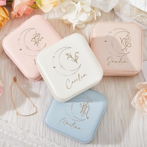 May include: Four square jewelry boxes in pastel colors: pink, white, and blue. Each box features a crescent moon design with a floral accent and a name. The boxes are surrounded by flowers and a gold necklace.