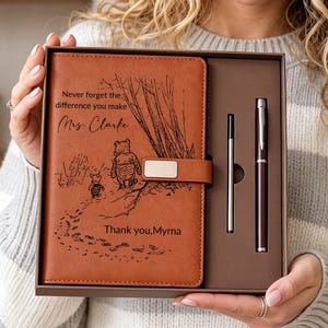 Könnte beinhalten: Ein beigefarbenes Notizbuch in Lederoptik mit Magnetverschluss, verziert mit einer schwarzen Strichzeichnung von Winnie Puuh und Ferkel, graviert mit dem Text "Never forget the difference you make Mrs. Clarke" und "Thank you, Myrna," inklusive passendem Stift.