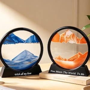 May include: Two circular sand art frames with black stands. The left frame contains blue sand forming mountain shapes with the text "With all my love" on the base. The right frame features orange sand forming mountain shapes with the text "You Mean The World To Me" on the base.