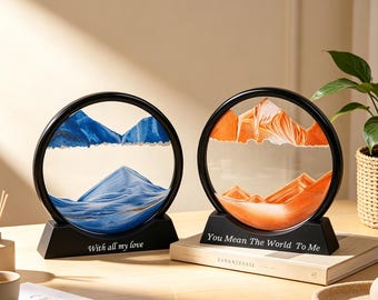 May include: Two circular sand art frames with black stands. The left frame contains blue sand forming mountain shapes with the text "With all my love" on the base. The right frame features orange sand forming mountain shapes with the text "You Mean The World To Me" on the base.
