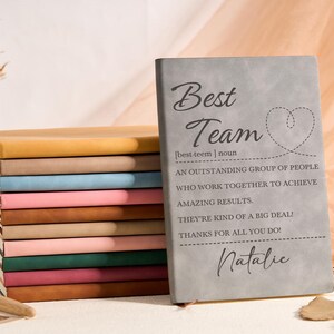 Puede incluir: Una pila de cuadernos coloridos con un cuaderno gris al frente. El cuaderno gris tiene las palabras "Best Team" y una definición impresa, junto con el nombre "Natalie". Hay un bolígrafo en la superficie.