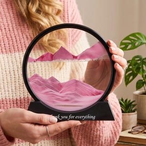 May include: A circular black frame containing pink sand that forms layered mountain shapes. The base of the frame is black and features the white text "Thank you for everything" printed on the front, creating a decorative sand art display.