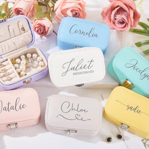 Może przedstawiać: Zestaw dziesięciu małych, prostokątnych pudełek na biżuterię w różnych pastelowych kolorach. Każde pudełko ma klapkę z zawiasem i mały zatrzask. Pokrywy są spersonalizowane nazwami, takimi jak "Natalie", "Chloe", "Jocelyn" i "Juliet Bridesmaid".