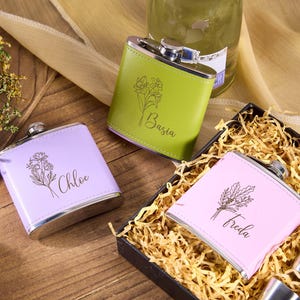 Puede incluir: Tres petacas personalizadas en lavanda, verde y rosa, cada una con un diseño floral y un nombre: Chloe, Basia y Freda. Las petacas se exhiben sobre una superficie de madera, con una caja de paja y vasos de chupito.