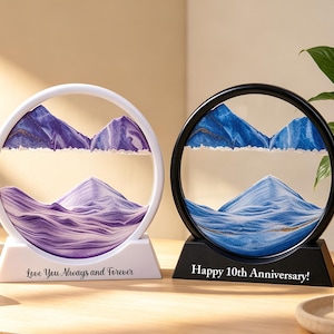 Könnte beinhalten: Zwei kreisförmige Sandkunst-Displays mit Sockeln. Der weiße Rahmen zeigt violetten Sand in Berglandschaften mit dem Text Love You Always and Forever. Der schwarze Rahmen zeigt blauen Sand in Berglandschaften mit dem Text Happy 10th Anniversary.