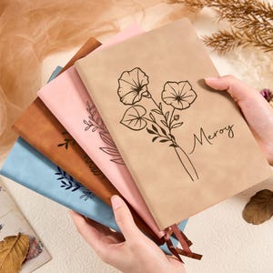 May include: A stack of notebooks in various colors, including tan, brown, pink, and blue. The tan notebook features a floral design and the word "Meroy". Each notebook has a ribbon bookmark.