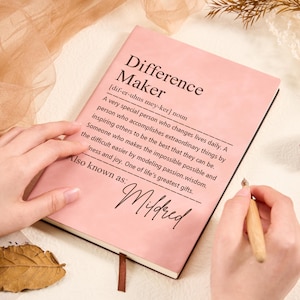 May include: A pink notebook with the words "Difference Maker" and a definition printed on the cover. A hand holds a pen, ready to write on the open notebook. The text also includes "Also known as: Mildred".