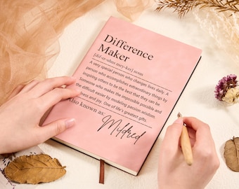 Könnte beinhalten: Ein rosa Notizbuch mit dem Text "Difference Maker" und einer Definition. Das Notizbuch ist geöffnet, eine Hand hält einen Stift, bereit zu schreiben. Der Text enthält auch "Also known as: Mildred".