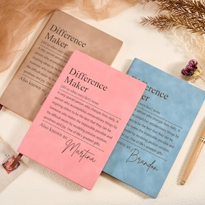 May include: Three notebooks in tan, pink, and blue, each with the definition of "Difference Maker" printed on the cover. The pink notebook is inscribed with the name "Martina", and the blue notebook has "Brandon". A gold pen and dried foliage are also present.