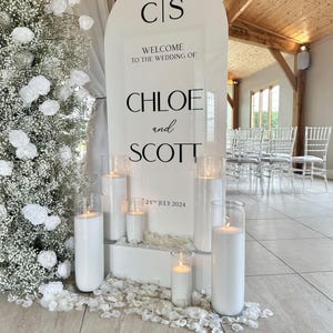 Op de afbeelding: Een wit welkomstschild voor een bruiloft met de tekst "CIS" bovenaan, gevolgd door "Welkom op de bruiloft van Chloe en Scott" en de datum "24 juli 2024". Het bord is versierd met witte kaarsen in glazen cilinders.