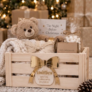 May include: A light-coloured wooden crate featuring a gold-coloured acrylic bow and the text "Hollie's SPECIAL CHRISTMAS DELIVERY". Inside are a tan teddy bear, a book titled "The Night Before Christmas", a bag of marshmallows, and hot chocolate.