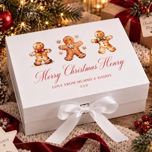 May include: A white rectangular gift box featuring three gingerbread person illustrations and the red printed text Merry Christmas Henry, Love From Mummy & Daddy XXX. The box is secured with a white satin ribbon tied in a bow at the front.