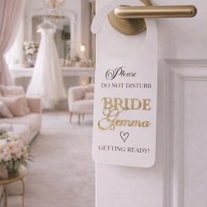 May include: White door hanger with gold text reading "Please DO NOT DISTURB BRIDE Gemma ♡ GETTING READY!". The door hanger is on a white door with a gold handle. A wedding dress is visible in the background, in a room with a chandelier.