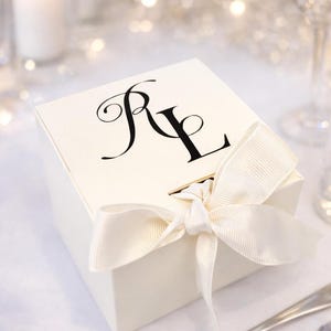 May include: A cream-coloured gift box tied with a matching satin ribbon. The box features the initials "RL" in a black, elegant script on the lid. The background is softly blurred with bokeh lights, suggesting a celebratory event.