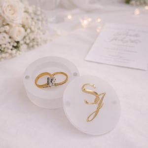 Peut inclure: Une bague de fiançailles en or avec une pierre centrale en diamant est présentée dans un écrin blanc. Le couvercle de la boîte porte des initiales dorées. La scène comprend des flûtes à champagne, des fleurs et une invitation de mariage.