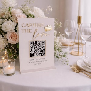 May include: A cream wedding sign with gold lettering saying "Capture the Love" and a QR code. A gold camera icon is above the word "love". The sign is on a white tablecloth, with flowers and candles in the background.