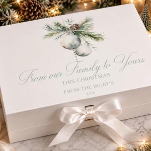 Könnte beinhalten: Eine weiße rechteckige Geschenkbox mit einer Aquarellillustration von Tannenzweigen und Weihnachtskugeln. Der Text lautet: From our Family to Yours, THIS CHRISTMAS, FROM THE RIGBY'S, xxx. Die Box ist mit einer weißen Satinschleife verschlossen.