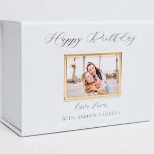 May include: White gift box with a gold frame around a photo of two women smiling. The text "Happy Birthday" is written in cursive on the box. The text "Love from Beth, Andrew & Harry" is written below the photo.