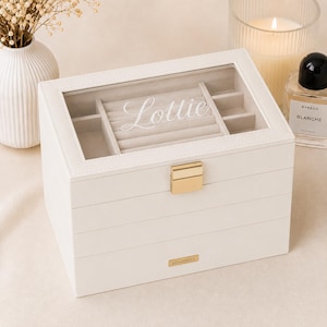 May include: A white rectangular jewellery box with a glass lid featuring the name Lottie in white script. The box has a gold-tone metal latch, a light grey velvet interior with ring rolls and compartments, and a small gold-tone Sohomics brand tag.