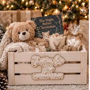 Könnte beinhalten: Eine helle Holzkiste mit der Aufschrift Christmas Eve Box FOR REGGIE & RUBY, die einen beigen Teddybären, ein Buch mit dem Titel The Night Before Christmas, eine Tasse mit Rentier-Motiv, eine Tüte mit weißen Marshmallows und eine Strickdecke enthält.