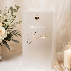 Pode incluir: Uma caixa retangular para cartões em acrílico transparente com um cadeado dourado em forma de coração no topo. A superfície frontal apresenta o texto branco MR & MRS e o nome Jones em letra cursiva dourada.