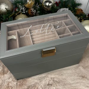 May include: A grey jewellery box with a clear glass top, featuring the name "Lottie" in cursive script. The box has multiple compartments and a gold-coloured clasp. The box is on a fluffy, light-coloured surface.