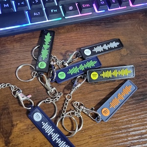 Könnte beinhalten: Eine Sammlung rechteckiger Schlüsselanhänger mit Spotify-ähnlichen Audiowellenmustern in verschiedenen Farben, darunter Schwarz, Blau und Grau. Jeder Schlüsselanhänger hat einen Metallverschluss und einen Ring für Schlüssel. Im Hintergrund befindet sich eine Tastatur.