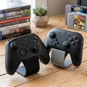 May include: Two black video game controllers resting on individual stands. The left controller features a directional pad, two analog sticks, and four colored action buttons. The right controller has a similar layout with a central logo and buttons.