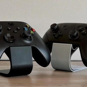 May include: Two black game controllers, one on a black stand and the other on a gray stand. The controllers have buttons in various colors. The stands have a curved design, providing a sleek display for gaming accessories.
