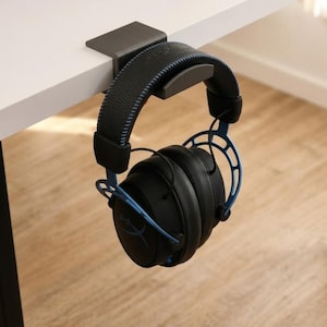 May include: A matte black metal headphone hanger mounted on the edge of a white desk, holding a pair of black over-ear headphones with blue metal accents, a textured headband, and a small red logo on the side of the ear cup.