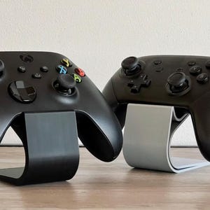 May include: Two black game controllers, one with colored buttons, rest on curved stands. The stand on the left is black, and the stand on the right is gray. The controllers are on a wooden surface.