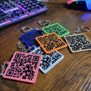 May include: A collection of colorful QR code keychains in pink, white, orange, blue, green, and tan. Each keychain features a black QR code design and a silver keyring. The keychains are arranged on a wooden surface.
