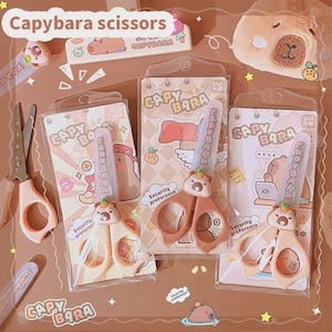 May include: A collection of capybara-themed scissors in various packaging. The scissors are peach-colored with capybara-shaped handles and protective covers. The packaging features capybara illustrations and the text "Capybara scissors".