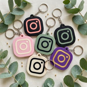 May include: A collection of six square keychains featuring the Instagram logo in various colour combinations, including black with coral, pink with white, sage green with black, black with sage green, cream with black, and purple with gold.