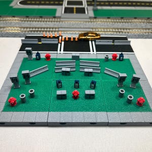 Op de afbeelding: Een miniatuur stadspark scène met een groen grasveld, grijze trottoirs en verschillende miniatuur gebouwen en objecten, waaronder brandkranen, lantaarnpalen en barrières.
