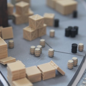Può includere: Modello in miniatura di un deposito ferroviario con vari elementi di carico. La scena include pile di pallet di legno, casse e barili nei toni del beige e del nero, disposti su una superficie grigia. I binari del treno modello sono visibili sullo sfondo.