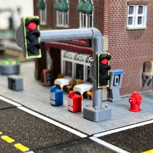 May include: A miniature traffic light set with two red lights, one on each side of a gray pole. The lights are yellow and have a black outline. The set is on a gray base and is part of a model city.