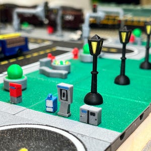 May include: A miniature city scene featuring a green park, a small truck, and four black streetlights with yellow lights. There are also several small buildings and a bench.