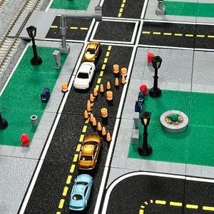 Puede incluir: Una escena de ciudad en miniatura con un atasco de tráfico de cuatro coches pequeños, un parque con una fuente y farolas. Los conos de tráfico naranja se colocan en una línea curva en la carretera.