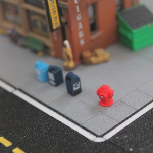 Op de afbeelding: Een miniatuur rode brandkraan, blauwe en zwarte brievenbussen en een groene vuilnisbak op een grijs en zwart straatbeeld.
