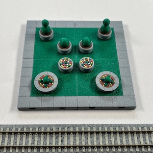 May include: A miniature model of a park with green grass, gray concrete paths, and colorful flower beds. There are small trees and bushes in the park.