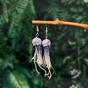 May include: A pair of handmade crochet jellyfish earrings. Each earring features a lavender-colored, crocheted bell shape with long, flowing white and lavender strands. The earrings hang from silver-colored hooks, displayed against a blurred green background.