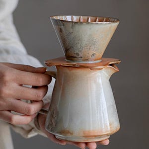 May include: A ceramic pour-over coffee maker set. The set features a cone-shaped dripper and a carafe with a handle and spout. The colours are a blend of white, brown, and orange, with a rustic, handmade appearance. The dripper sits atop a flat, circular base.